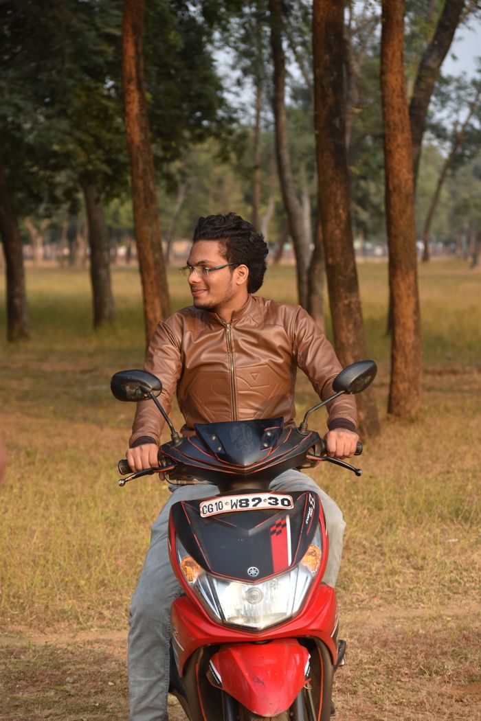 A man wearing a leather jacket sits on a red motorcycle in a park with trees.