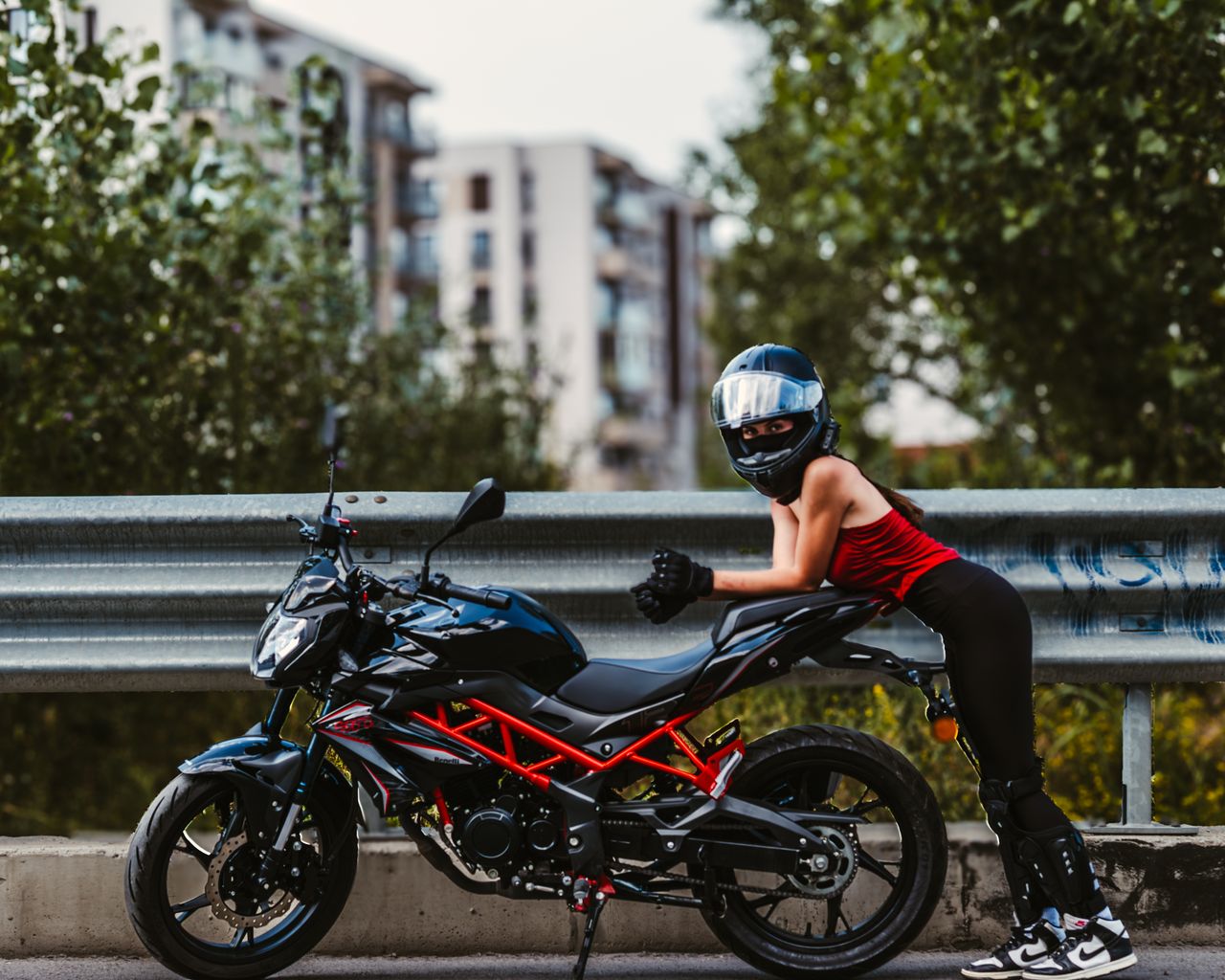 Female motorcycle rider in gear poses beside bike on urban roadside, showcasing modern biking culture.