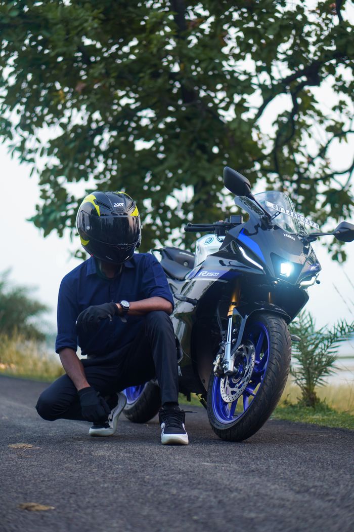 A person wearing a helmet crouching beside a sports motorcycle on a scenic road.