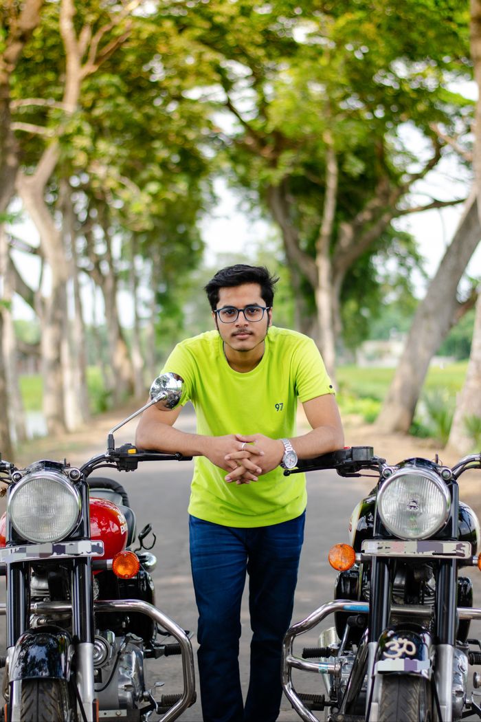 Man in glasses poses with vintage motorcycles on a scenic road, showcasing style and confidence.